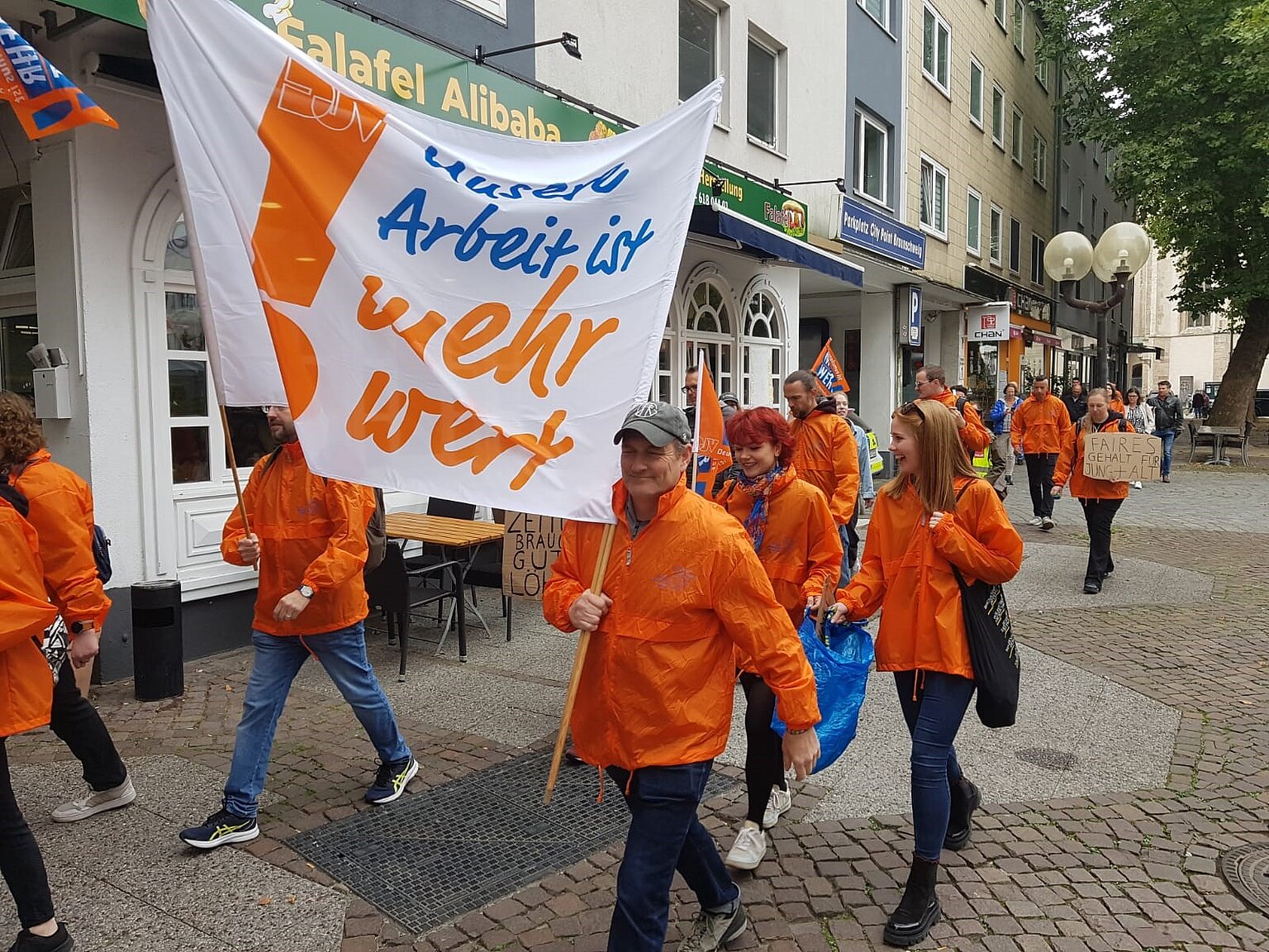 Menschen mit oragefarbigen Jacken laufen mit Demonstrationsplakat durch eine Straße – 